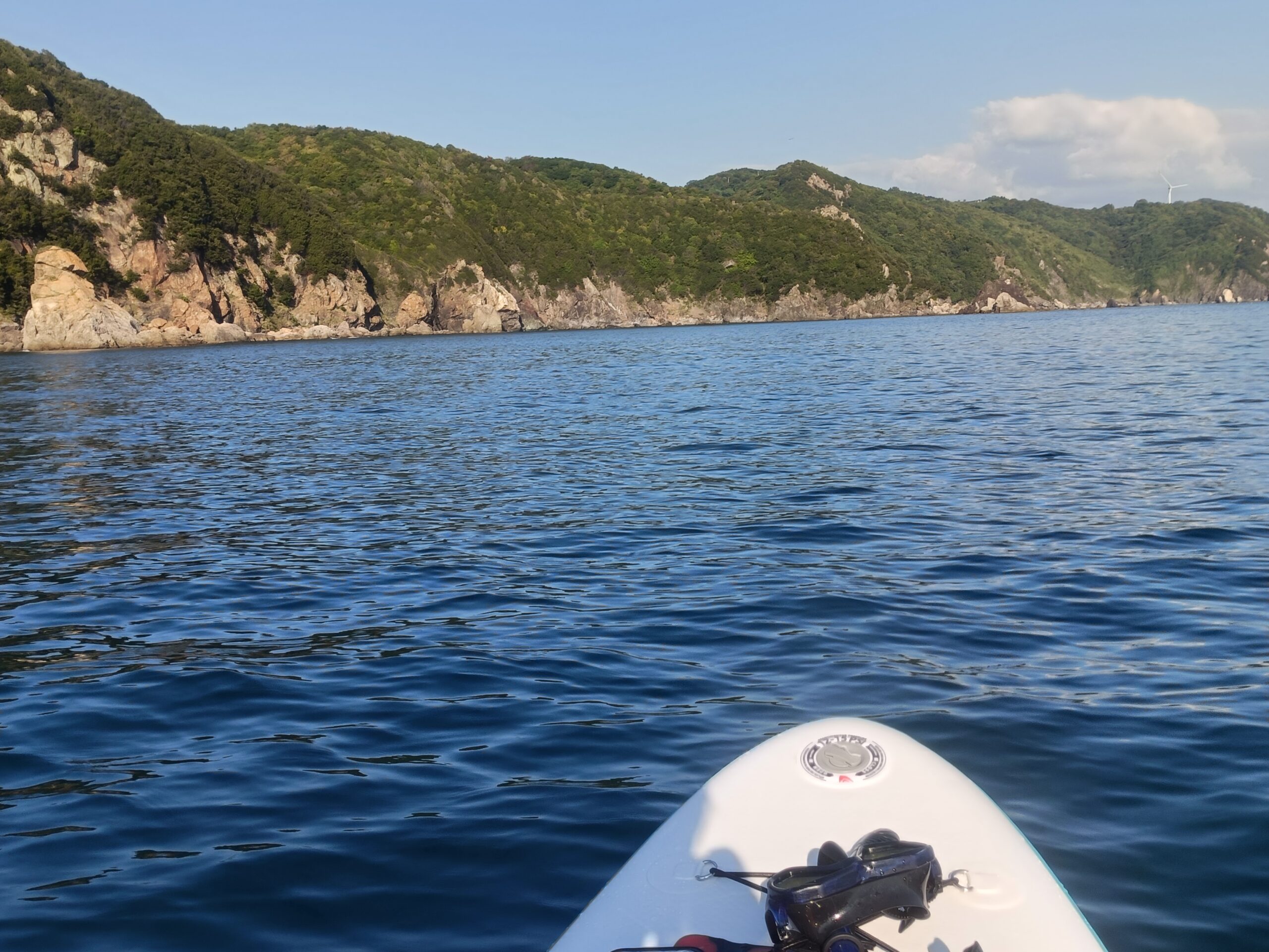 SUPのボードの先端越しに望む青い海と、緑に覆われた岩の海岸線