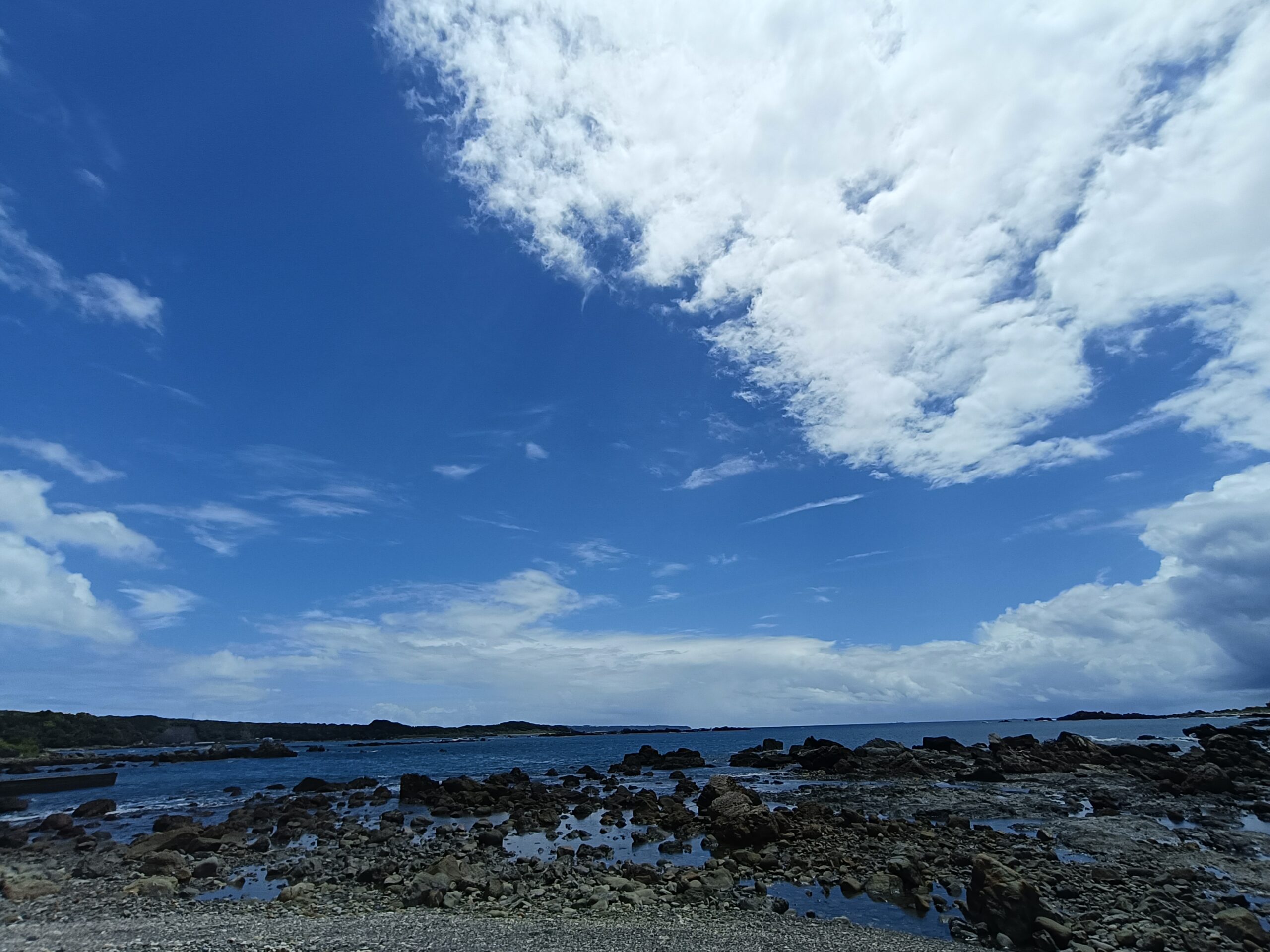 青空と白い雲の下に潮だまりのある岩礁と穏やかな海が広がる
